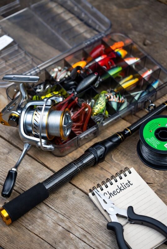 A flat lay of essential fishing gear including a reel, rod, lures, and a checklist.